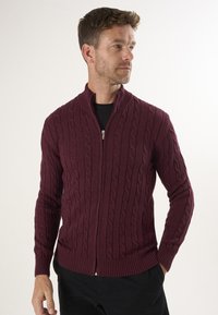 Burgunderfarbener gestrickter Reißverschluss-Cardigan mit einem Kabelstrickmuster, gerippten Bündchen und einem hohen Kragen für einen taillierten Look.