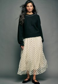 Czarny sweter dzianinowy z długimi rękawami, nałożony na kremową, przezroczystą maxi spódnicę w kropki. Czarną parę balerin dopełnia outfit.