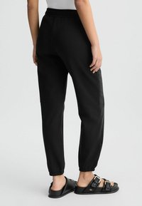 Pantalons de jogging noirs avec une taille élastique, des jambes fuselées et des panneaux latéraux en maille, associés à des sandales noires à glisser avec doubles boucles de fermeture.