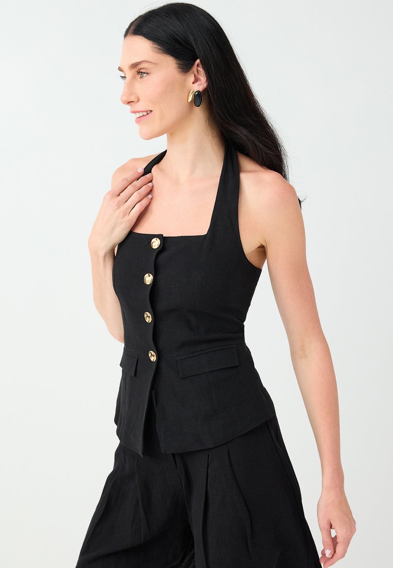 Femme aux longs cheveux foncés porte un haut noir sans manches col halter avec des boutons dorés et un pantalon noir assorti, souriante et regardant à gauche.