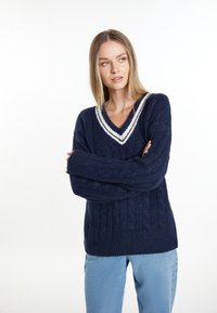 Marineblauer Zopfmuster-Pullover mit V-Ausschnitt und weißen Streifenakzenten. Verfügt über lange Ärmel und eine lockere Passform. Getragen über hellblauen Jeans.