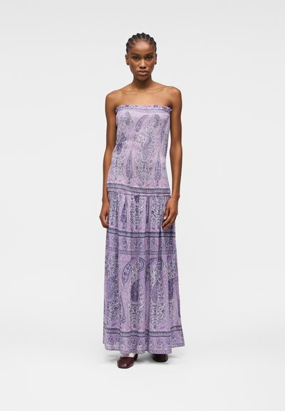 Femme portant une robe violette sans bretelles, longue jusqu'au sol, avec un motif cachemire, debout les bras détendus le long du corps devant un fond uni.
