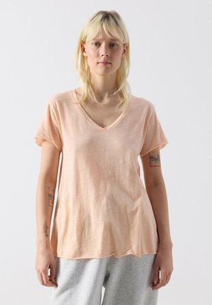 Junge Frau mit blonden Haaren, die ein lockeres, pfirsichfarbenes V-Ausschnitt-T-Shirt und graue Hose trägt, steht vor einem schlichten hellen Hintergrund.