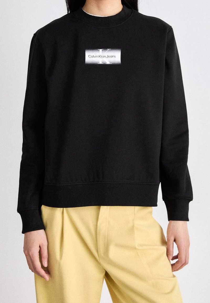 Svart sweatshirt med rund halsringning, ribbade manschetter och nederkant. Har en vit logotyp och texten "Calvin Klein Jeans" centrerad på framsidan.
