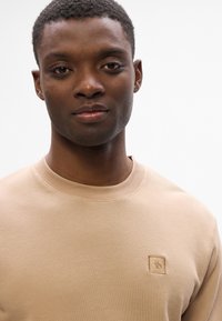 Primo piano di un uomo che indossa un maglione beige a collo rotondo con un piccolo logo ricamato sul petto, che sorride leggermente su uno sfondo bianco.