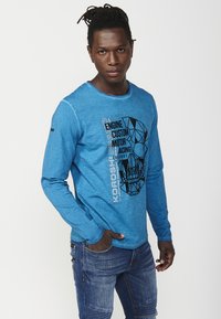 Blue long-sleeve shirt made of textured fabric, featuring a black graphic design of a skull and text, with a round neckline and stitched details.