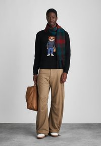 Schwarzer Pullover mit einem gestrickten Bären-Graphic, beigen weiten Hosen und einem karierter Schal in Dunkelgrün, Rot und Schwarz. Braune Tote-Tasche. Weiße Schuhe.