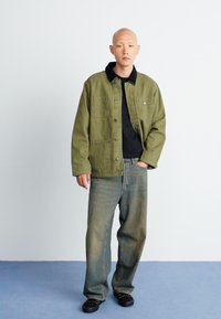 Olivengrøn jakke med to forlommer, sort krave, båret over en sort skjorte. Kombineret med blågrå jeans og sorte sko.