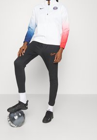 Chaqueta blanca con mangas de degradado de azul a rojo, logo de París, pantalones de chándal negros con logo naranja de Nike, y zapatos de fútbol negros sobre un balón negro.