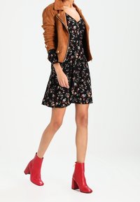 Veste en daim marron portée sur une robe noire à fleurs. La robe a un corsage ajusté et une jupe évasée. Des bottines rouges à talons carrés complètent le look.
