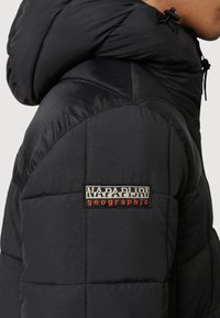 Osoba w czarnej pikowanej kurtce z kapturem, z prostokątną naszywką z logo Napapijri Geographic na górnej części ramienia.