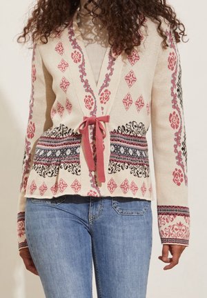 Femme portant un cardigan crème avec des motifs floraux et géométriques roses, noirs et bleus, noué avec un ruban rose, associé à un jean bleu.