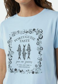 Jasnoniebieski t-shirt z krótkimi rękawami z czarnym graficznym wzorem przedstawiającym ryby i elementy florystyczne, z napisem "Portugalski Smak" na środku.
