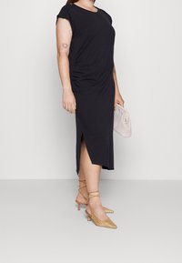 Černé šaty po kolena s krátkými rukávy a bočním rozparkem, doplněné béžovými kostkovanými lodičkami s špičatou špičkou a texturovanou béžovou kabelkou.