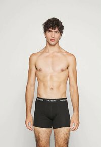 Fiatal férfi göndör hajjal áll szemben, fekete boxer alsóneműt visel, amelynek derékrészén az "ONLY&SONS" logó található.