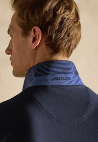 Tumesinine polo särk sinise triibuga kraega, millel on seljal "JOULES" ära õmmeldud. Sujuv tekstuur, keha lähedane disain ja klassikaline lõige.