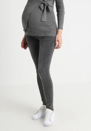 Schwangere Person trägt einen grauen gerippten Pullover mit gebundener Taille, dunkelgraue Skinny Jeans und weiße Sneaker, stehend vor hellem Hintergrund.
