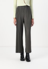 Persona che indossa pantaloni grigi a gamba larga e vita alta con pieghe, un maglione a coste giallo chiaro e stivali neri con tacco alla caviglia, in piedi davanti a uno sfondo bianco.