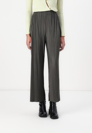 Personne portant un pantalon gris plissé à taille haute et jambes larges, un pull en maille côtelée jaune clair et des bottines à talons noires, se tenant contre un fond blanc.