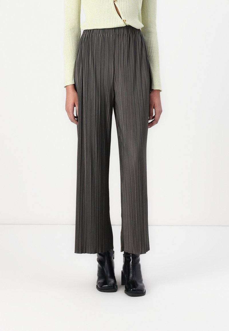 Persona che indossa pantaloni grigi a gamba larga e vita alta con pieghe, un maglione a coste giallo chiaro e stivali neri con tacco alla caviglia, in piedi davanti a uno sfondo bianco.