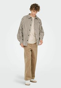 Geruite overhemd in bruintinten, gedragen over een beige T-shirt, met losse corduroy broek in beige en witte sneakers.