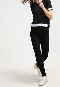 Žena s kudrnatými vlasy má na sobě černý top s krátkým rukávem a vrstveným designem, černé úzké džíny a bílé tenisky, stojí s jednou nohou překříženou.