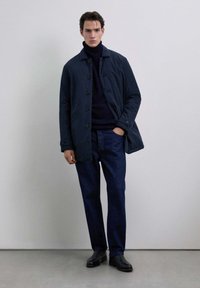 Hombre con chaqueta azul marino oscuro, suéter de cuello alto negro, jeans azul oscuro y botas negras, de pie frente a un fondo gris claro liso.