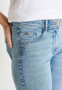Jeans de mezclilla azul claros con un corte ajustado, que cuentan con un diseño tradicional de cinco bolsillos, costuras sutiles y un botón y remaches de metal.