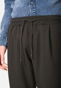 Pantaloni jogger neri con vita elasticizzata e dettaglio con cordino. Tessuto liscio con pieghe e una leggera lucentezza. Abbinati a una camicia di jeans.