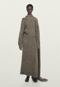 Modella di colore che indossa un lungo vestito grigio a maglia texture con una sciarpa attaccata che scende su una spalla e zoccoli neri.