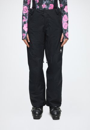 Personne portant un pantalon de neige noir avec des bretelles et des bottes de ski noires, associé à un haut à manches longues fleuri.
