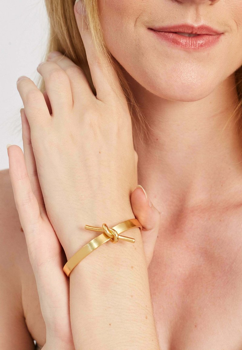 Zlatý kovový náramok s hladkým pásom a akcentom uzla. Dizajn je minimalistický s lesklým povrchom a jednoduchým, čistým tvarom.