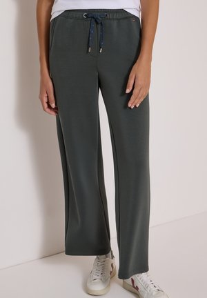 Personne portant un pantalon large vert foncé avec une taille à cordon et des baskets blanches, debout devant un fond clair uni.