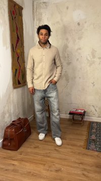 Junger Mann mit Dreadlocks in beigem Hemd und grauer Jeans steht drinnen neben einer Ledertasche und einem kleinen Hocker mit Büchern auf dem Holzboden.