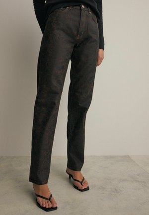 Tummanruskeat kuviolliset farkut suoran leikkauksen omaavina, joissa on kuparinväriset ompeleet ja klassinen viiden taskun muotoilu, yhdistettynä mustiin hihnaisiiin sandaaleihin.