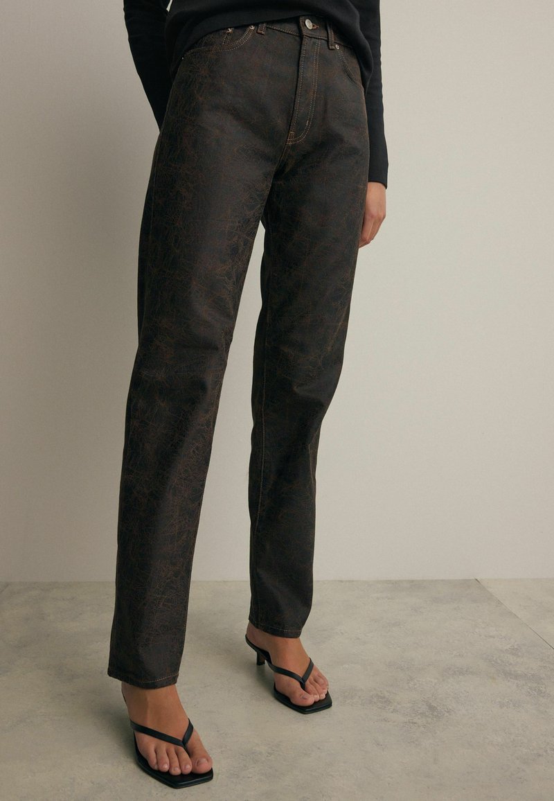 Jeans de patrón marrón oscuro con corte recto, que presentan costuras en color cobre y un diseño clásico de cinco bolsillos, combinados con sandalias negras de tiras.