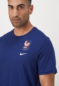 Nike mėlynas marškinėliai su trumpomis rankovėmis, turintis rožinį gaidžio emblema ir "FFF" ant krūtinės. Lygus audinys, apvalaus kaklo dizainas.