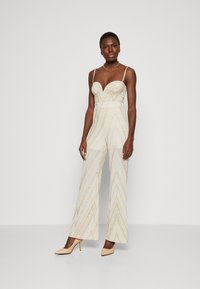 Beige Jumpsuit mit Herzausschnitt, schmalen Trägern und einem Chevron-Muster mit goldenen Akzenten. Weite Beinpartie; weicher, strukturierter Stoff.