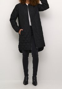 Frau in einem langen schwarzen gesteppte Mantel, schwarzen Skinny-Jeans, schwarzen Stiefeletten und einem weißen Hemd, stehend vor einem schlichten Hintergrund.