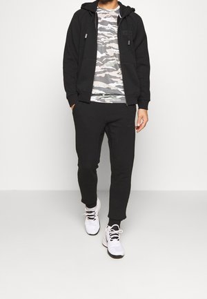 Schwarzer Kapuzenpullover mit Zugkordeln, Camo-T-Shirt, schwarze Jogginghose und weiße Sneakers mit schwarzen Sohlen. Mit Logo auf dem Hoodie und der Hose.