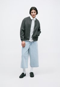 Grijze bomberjack, lichtgrijs polo-shirt, brede lichtblauwe denim culottes, witte sokken en zwarte instappers. Gladde texturen en moderne pasvorm.