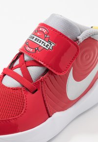 Červená atletická bota s lesklým povrchem, šedou síťovanou jazyku a stříbrným logem Nike. Obsahuje suchý zip a texturované detaily na boku.