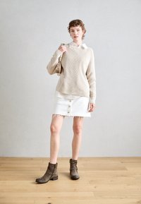 Suéter beige de canalé, falda de mezclilla blanca con detalles de botones y botines grises. La modelo sostiene un bolso pequeño, con un fondo neutro.