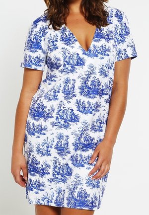 Frau trägt ein weißes Kleid mit blauem Landschafts-Toile-Druck, kurzen Ärmeln und V-Ausschnitt, steht vor einfachem Hintergrund.