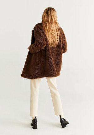 Kobieta z długimi blond włosami, ubrana w brązowy, teksturowany płaszcz, kremowe spodnie i czarne buty na obcasie, stojąca tyłem.