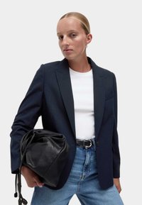 Blazer marine avec revers crantés, associé à une chemise blanche et un jean bleu taille haute. Tenue d'un sac en cuir noir à cordon.