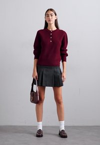 Maglione ribbed bordeaux con tre bottoni, gonna piegata grigia, calzini bianchi e mocassini neri con borchie decorative su uno sfondo neutro.