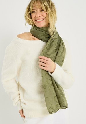 Mujer sonriente con cabello rubio que lleva un suéter blanco de hombros descubiertos y sostiene una bufanda estampada verde oliva envuelta alrededor de su cuello.