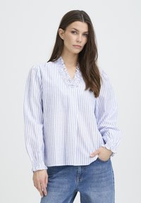 Camicia bianca con righe verticali blu, scollo a V con volant e polsini elasticizzati. Il materiale sembra leggero e strutturato. Abbinata a denim.