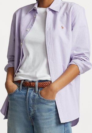 Persoon draagt een lichtpaarse overhemd met knopen over een wit T-shirt, blauwe jeans met een gevlochten bruine riem, handen in de zakken.